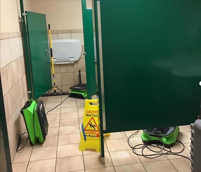 Three SERVPRO air movers and a dehumidifier operating in a commercial restroom with green stalls, tile floor, and a wet floor